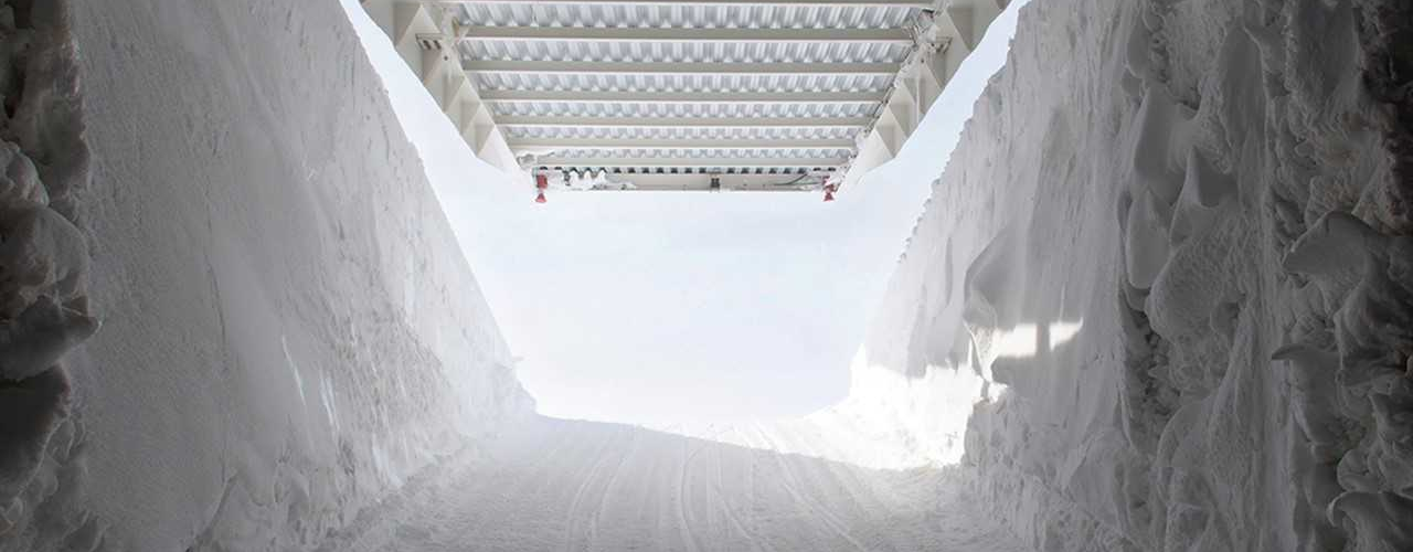 Das Bild zeigt das Innere eines Tunnels aus Eis und Schnee. Er führt auf einer Rampe aus festgetretenem Schnee hinaus in die Sonne und in eisblauen Himmel. Dabei hebt sich das weiße Stahldach über dem Eistunnel und gibt das helle Sonnenlicht frei.