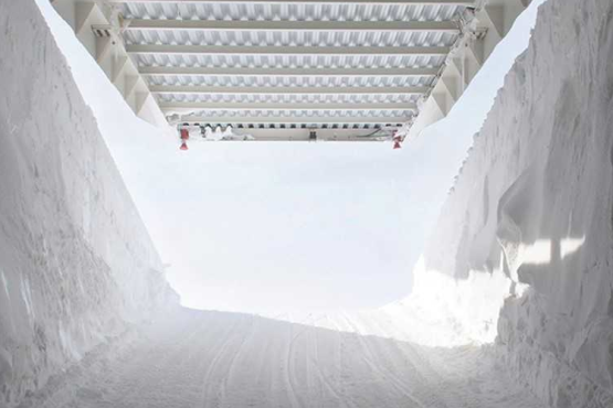 The picture shows the inside of a tunnel made of ice and snow. It leads out on a ramp of packed snow into the sun and an ice-blue sky. The white steel roof above the ice tunnel rises and reveals the bright sunlight.