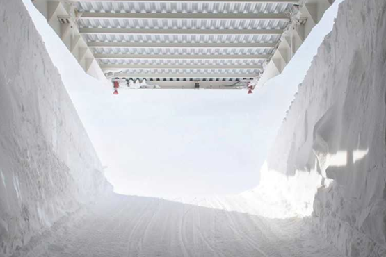 Das Bild zeigt das Innere eines Tunnels aus Eis und Schnee. Er führt auf einer Rampe aus festgetretenem Schnee hinaus in die Sonne und in eisblauen Himmel. Dabei hebt sich das weiße Stahldach über dem Eistunnel und gibt das helle Sonnenlicht frei.