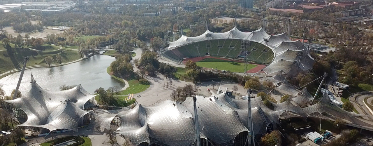 Man sieht ein Luftbild des Münchner Olympiaparks, mit dem Olympiastadion und seinem weltberühmten Zeltdach und dem Olympiasee