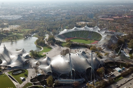 Man sieht ein Luftbild des Münchner Olympiaparks, mit dem Olympiastadion und seinem weltberühmten Zeltdach und dem Olympiasee
