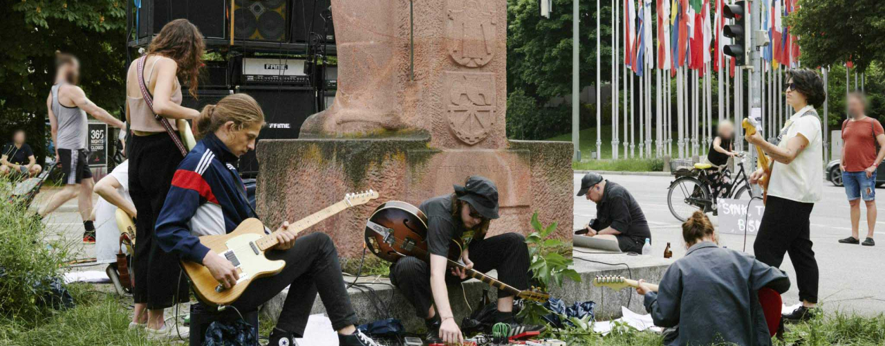 Junge, leger gekleidete Menschen, die mit umgeschnallten E-Gitarren rund um das Bismarck-Denkmal im Stehen und Sitzen und musizieren. Hinter dem Denkmal ist eine Wand aus Gitarrenverstärkern aufgebaut.