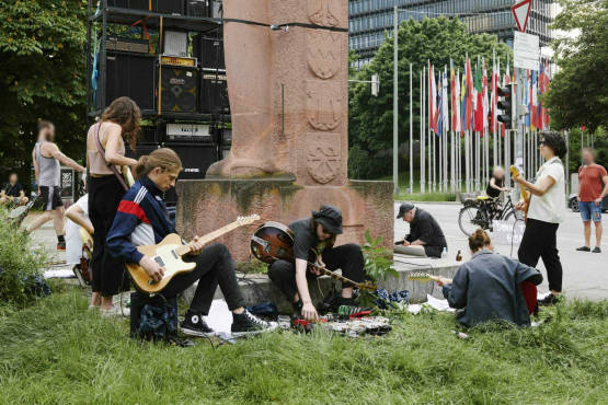 Junge, leger gekleidete Menschen, die mit umgeschnallten E-Gitarren rund um das Bismarck-Denkmal im Stehen und Sitzen und musizieren. Hinter dem Denkmal ist eine Wand aus Gitarrenverstärkern aufgebaut.