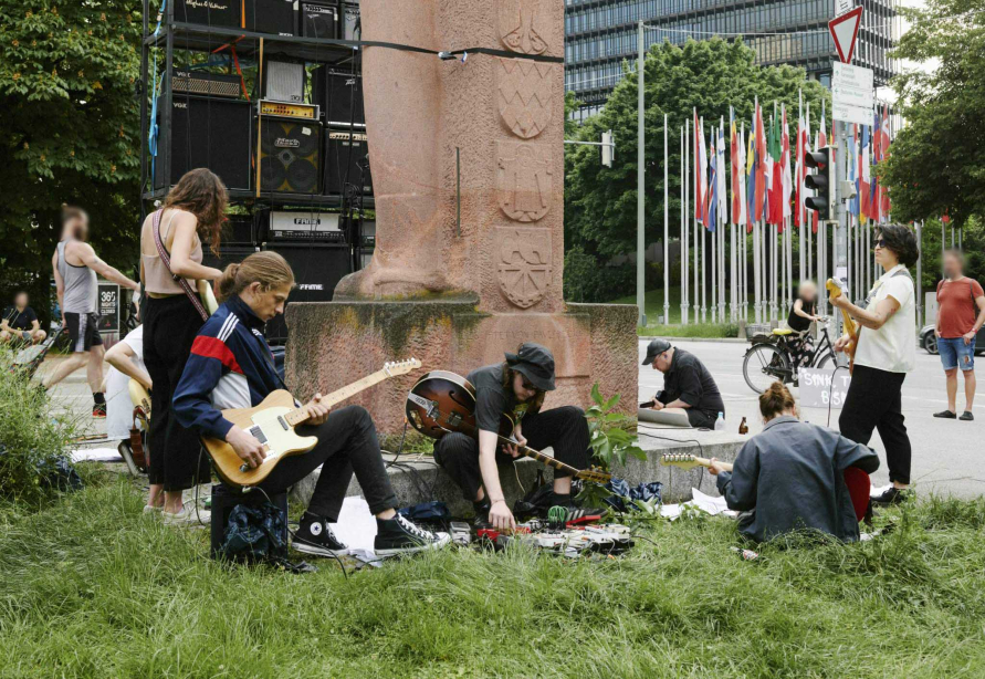 Young, casually dressed people standing and sitting around the Bismarck monument with their electric guitars on and making music. A wall of guitar amps is set up behind the monument.
