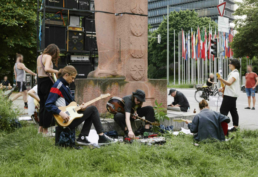 Junge, leger gekleidete Menschen, die mit umgeschnallten E-Gitarren rund um das Bismarck-Denkmal im Stehen und Sitzen und musizieren. Hinter dem Denkmal ist eine Wand aus Gitarrenverstärkern aufgebaut.