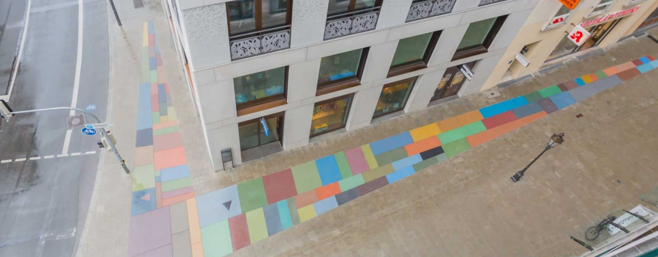 Aerial perspective from above looking down on a house facade and the wide sidewalk at a street crossing. A 70 square meter floor mosaic made of colored stones in the form of a large angle is embedded in the pavement of the sidewalk.