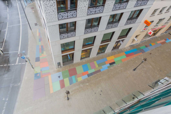 Aerial perspective from above looking down on a house facade and the wide sidewalk at a street crossing. A 70 square meter floor mosaic made of colored stones in the form of a large angle is embedded in the pavement of the sidewalk.