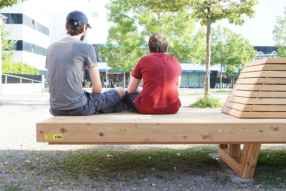 Two young people sitting on the wooden bench together, photographed from behind. The mira shopping center at Nordhaideplatz can be seen in the background.