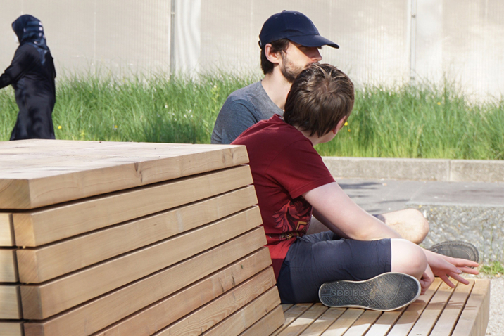 Two teenagers talking, sitting on the wooden bench. In the background passers-by walk.