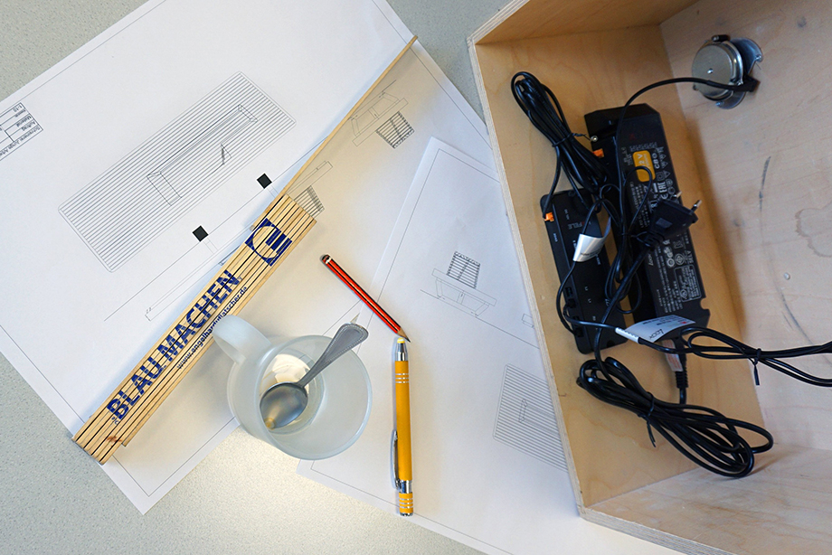 Table with construction drawings for the bench, pens, a cup and a meter rule, photographed from above. Next to it you can see a wooden box with built-in electronic components.