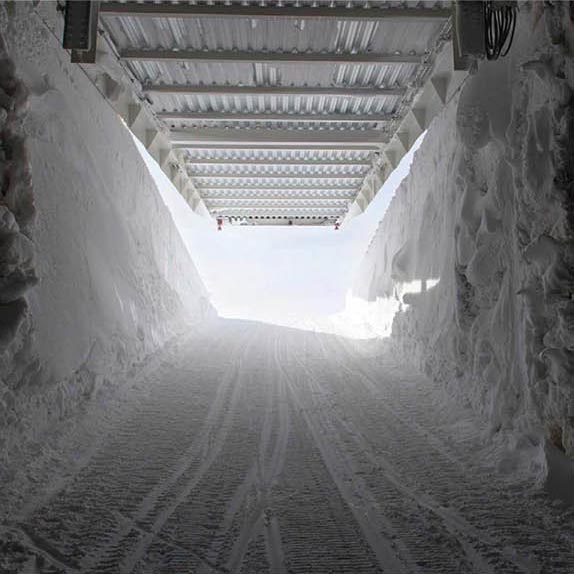 The picture shows the inside of a tunnel made of ice and snow. It leads out on a ramp of packed snow into the sun and an ice-blue sky. The white steel roof above the ice tunnel rises and reveals the bright sunlight.