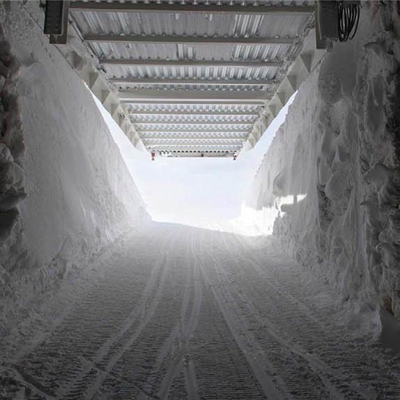 Das Bild zeigt das Innere eines Tunnels aus Eis und Schnee. Er führt auf einer Rampe aus festgetretenem Schnee hinaus in die Sonne und in eisblauen Himmel. Dabei hebt sich das weiße Stahldach über dem Eistunnel und gibt das helle Sonnenlicht frei.