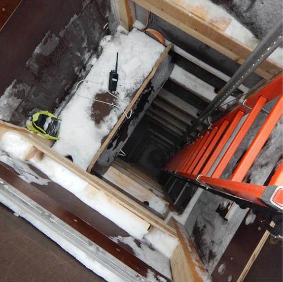 Man sieht auf einer Baustelle von oben in einen tiefen Schacht hinein, konstruiert aus Holz. In den Schacht hinunter führt eine rote Leiter. Oben am Rand des Schachts befindet sich Eis und Schnee; ein Funkgerät liegt am Schachteingang.