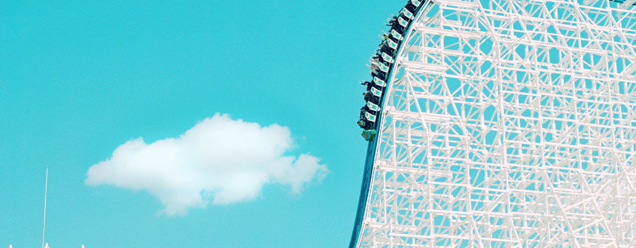 Against the background of the blue sky you can see the white structure of a roller coaster, which in this section drops steeply from top right to bottom left and is being ridden by an almost full car.