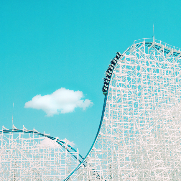 Against the background of the blue sky you can see the white structure of a roller coaster, which in this section drops steeply from top right to bottom left and is being ridden by an almost full car.