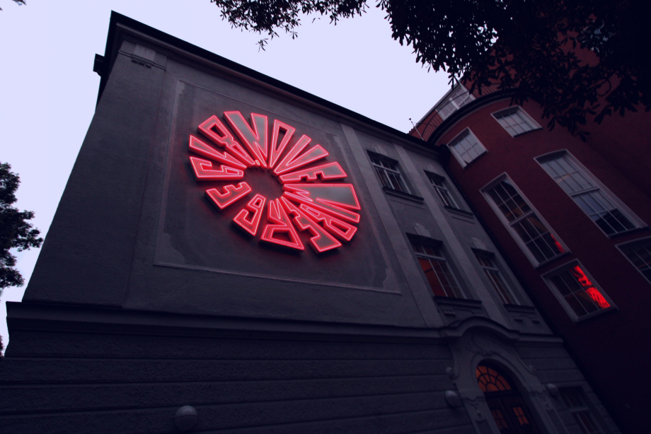 At dusk, an installation of bright red neon tubes appears on a house wall in the shape of a bomb detonation. The diameter of the installation is about 5 meters.