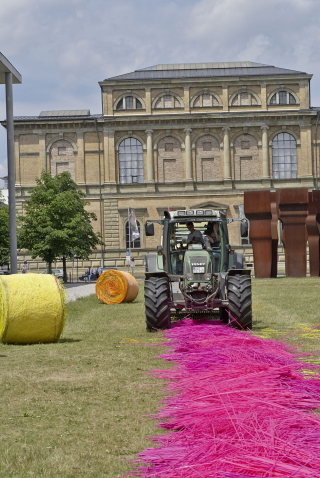 A tractor in a meadow presses long pink straws into bales. In the background the Alte Pinakothek and a yellow and an orange wrapped bale.