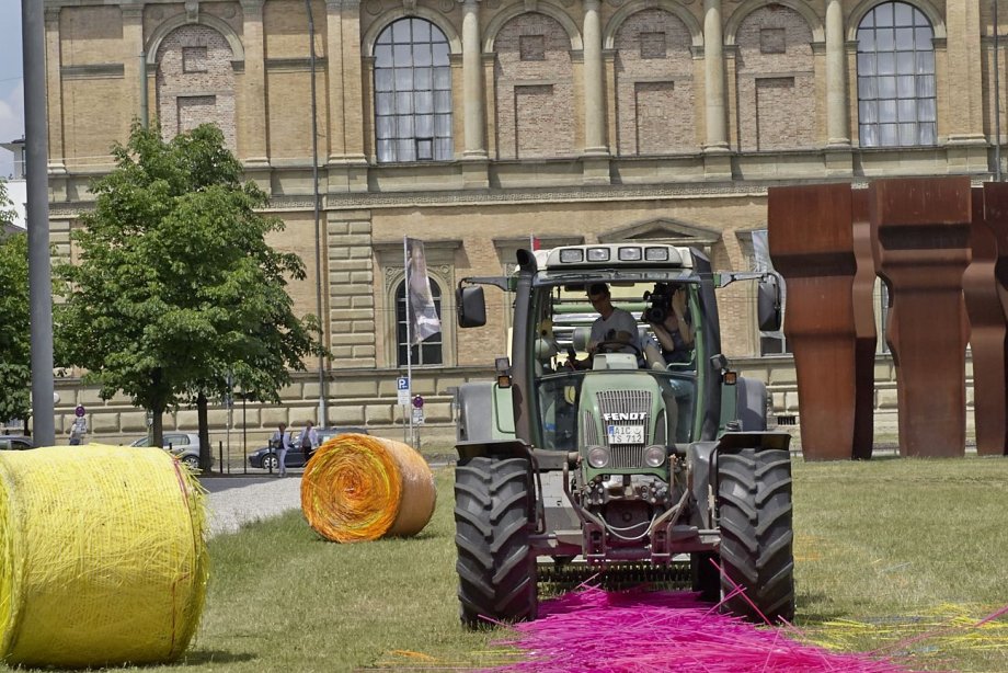 A tractor in a meadow presses long pink straws into bales. In the background the Alte Pinakothek and a yellow and an orange wrapped bale.