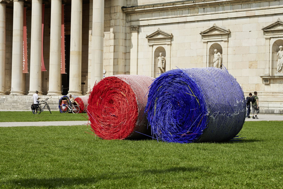 On the meadow in front of the Glyptothek on Königsplatz, in the foreground of the picture, lies a large red and blue bale of straw. In the background you can see a woman with a bicycle posing for a photo in front of two more bales.