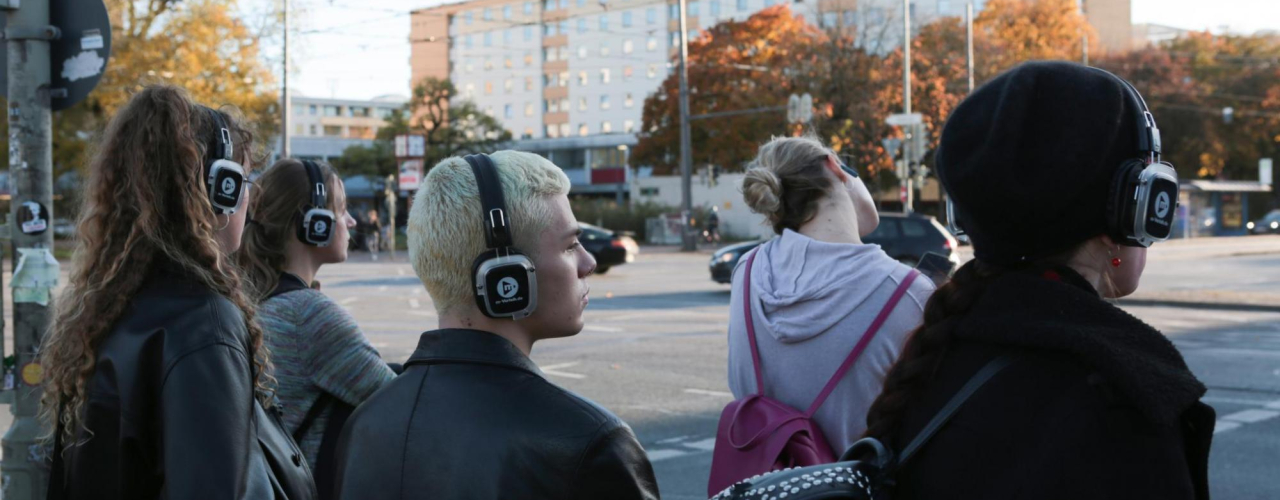 Five young people with headphones are standing in front of a pedestrian traffic light in city traffic