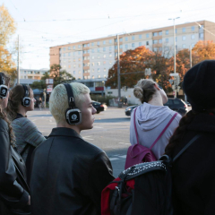 Fünf Jugendliche mit Kopfhörern stehen vor einer Fussgängerampel im Stadtverkehr