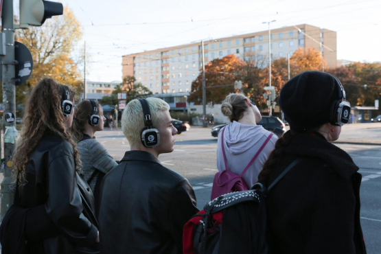 Fünf Jugendliche mit Kopfhörern stehen vor einer Fussgängerampel im Stadtverkehr