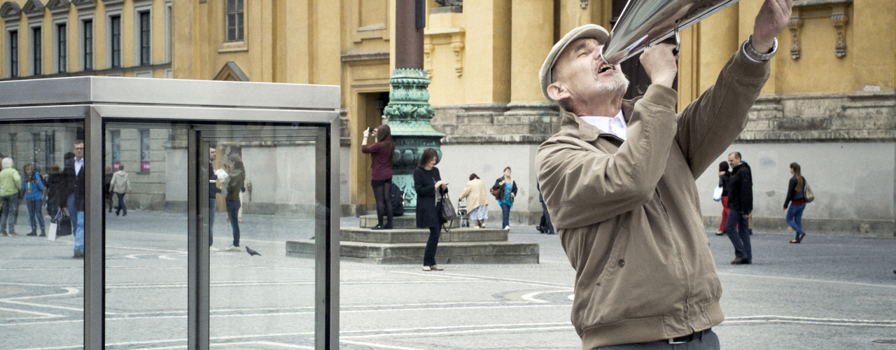 An older man with a brown jacket, gray trousers and a peaked cap is standing on the Odeonsplatz next to a man-high glass display case. He holds a large megaphone above his head and shouts loudly into it. In the background you can see passers-by on the square.