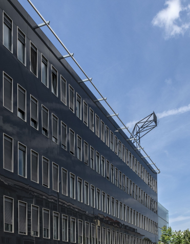 You can see the high and closed black facade of a five-story house with long, exactly the same size window fronts on each floor. A basketball hoop wall is installed on top of the roof. She rises up into the blue sky.