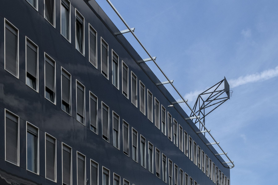 You can see the high and closed black facade of a five-story house with long, exactly the same size window fronts on each floor. A basketball hoop wall is installed on top of the roof. She rises up into the blue sky.
