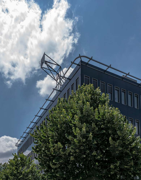 Auf dem Dach eines fünfstöckigen hohen Hauses ist eine Basketballkorbwand angebracht. Der Blick geht von der Straße unten in die Höhe, über der Basketballkorbwand auf dem Dach spannt sich weiß-blauer Sommerhimmel. Es erscheint absurd, in dieser Höhe einen Ball in den Korb zu treffen.