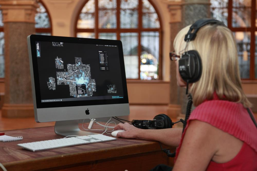 A woman sits in front of a computer with headphones on. The memoryloops.net website is open on the screen. The woman uses the mouse cursor to search for different audio tracks and listens to them.