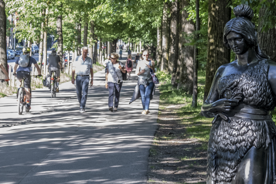 Im sommerlichen München sieht man drei Radfahrer auf einem Radweg, rechts daneben am Bürgersteig gehen drei Passanten. Ganz rechts im Vordergrund steht eine ca 160cm große Bronzestatue der Bavaria. Ohne die kriegerischen Attribute Löwe und Schwert ist sie auf Augenhöhe mit den Passant*innen dargestellt.