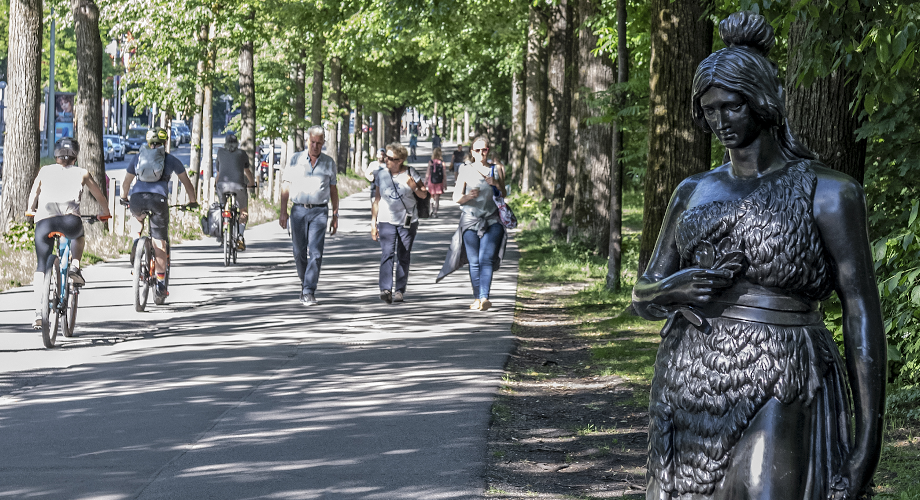 In summertime Munich you can see three cyclists on a bike path, three passers-by are walking on the sidewalk to the right. On the far right in the foreground is a 160cm tall bronze statue of Bavaria. Without the martial attributes of lion and sword, she is shown at eye level with passers-by.
