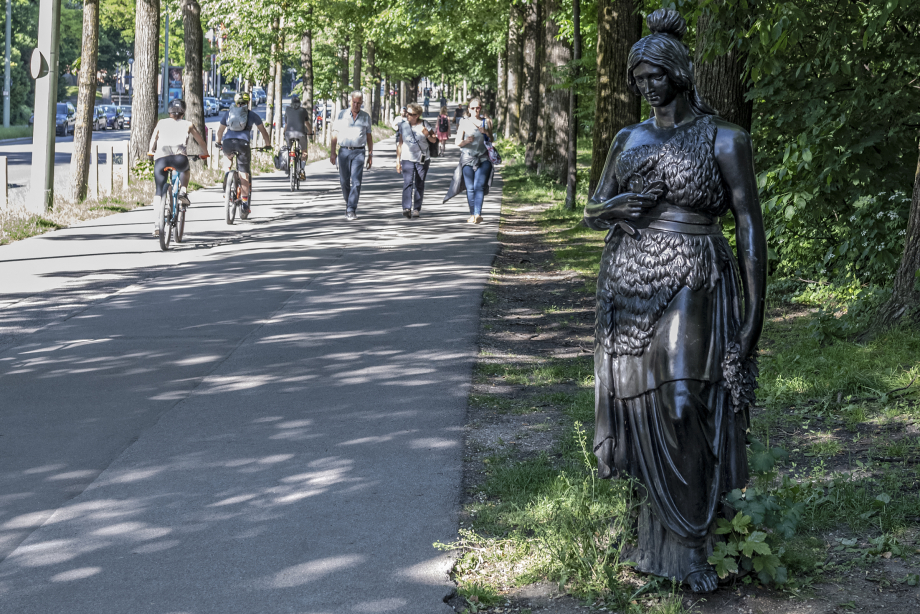 In summertime Munich you can see three cyclists on a bike path, three passers-by are walking on the sidewalk to the right. On the far right in the foreground is a 160cm tall bronze statue of Bavaria. Without the martial attributes of lion and sword, she is shown at eye level with passers-by.