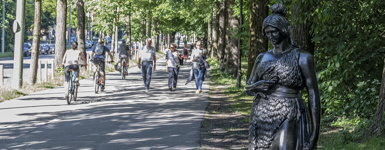 In summertime Munich you can see three cyclists on a bike path, three passers-by are walking on the sidewalk to the right. On the far right in the foreground is a 160cm tall bronze statue of Bavaria. Without the martial attributes of lion and sword, she is shown at eye level with passers-by.