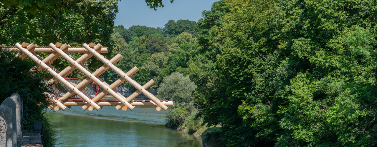 Man sieht einen hölzernen Brückenteil, der nur bis zur Hälfte über die Isar ragt. Darunter fliesst die grüne Isar, umrahmt von dicht belaubten Bäumen an einem Sommertag.