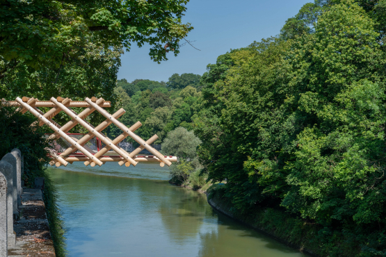 Man sieht einen hölzernen Brückenteil, der nur bis zur Hälfte über die Isar ragt. Darunter fliesst die grüne Isar, umrahmt von dicht belaubten Bäumen an einem Sommertag.