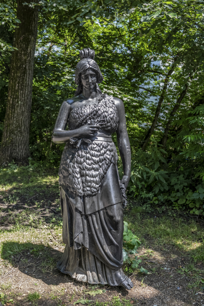 The picture shows a bronze sculpture of Bavaria facing the viewer. The female figure is shown without sword and lion and stands gracefully on the banks of the Isar. In the background are trees. The sun is shining.
