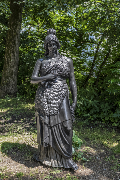 Das Bild zeigt eine Bronzeplastik der Bavaria, die den Betrachtenden zugewandt ist. Die Frauengestalt wird ohne Schwert und Löwe dargestellt und steht anmutig am Isarufer. Im Hintergrund sind Bäume. Die Sonne scheint.