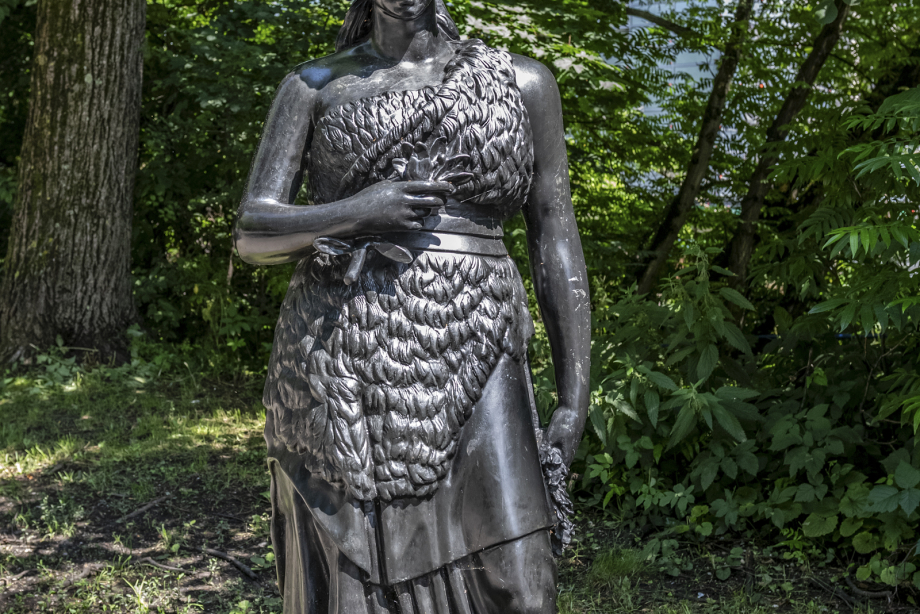 The picture shows a bronze sculpture of Bavaria facing the viewer. The female figure is shown without sword and lion and stands gracefully on the banks of the Isar. In the background are trees. The sun is shining.