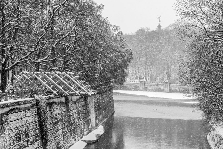 Ein hölzerner Brückenteil ragt über die Isar. Es ist Winter, Schnee liegt auf dem Brückenteil, unten am Isarufer und auf den Bäumen an beiden Uferseiten. Im Hintergrund sieht man den Friedensengel