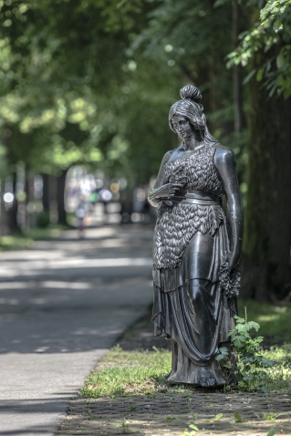 Die Bronzeplastik der Bavaria steht anmutig und auf menschliches Maß herunterskaliert vor dem Hintergrund von Bäumen direkt neben einem Bürgersteig.