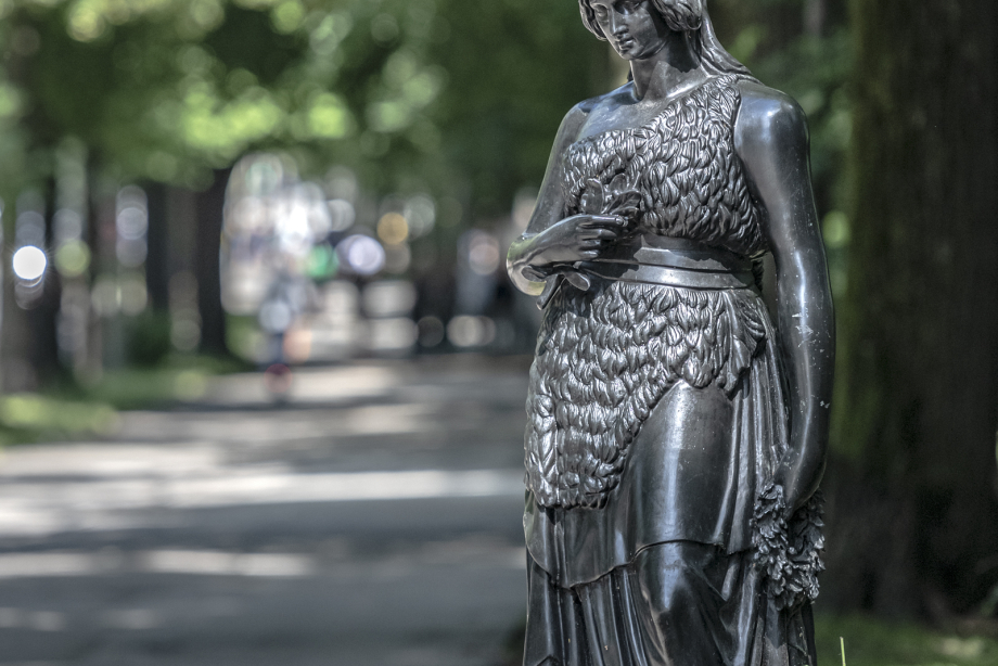 Die Bronzeplastik der Bavaria steht anmutig und auf menschliches Maß herunterskaliert vor dem Hintergrund von Bäumen direkt neben einem Bürgersteig.