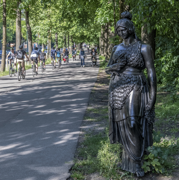 Die lebensgroße Bronzeplastik der Bavaria steht rechts im Bildvordergrund. Daneben sieht man Menschen am Bürgersteig und Radler*innen am Radfahrweg.