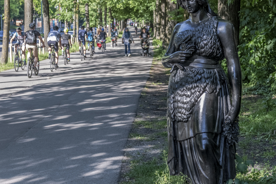 The life-size bronze sculpture of Bavaria is in the foreground on the right. You can also see people on the sidewalk and cyclists on the cycle path.
