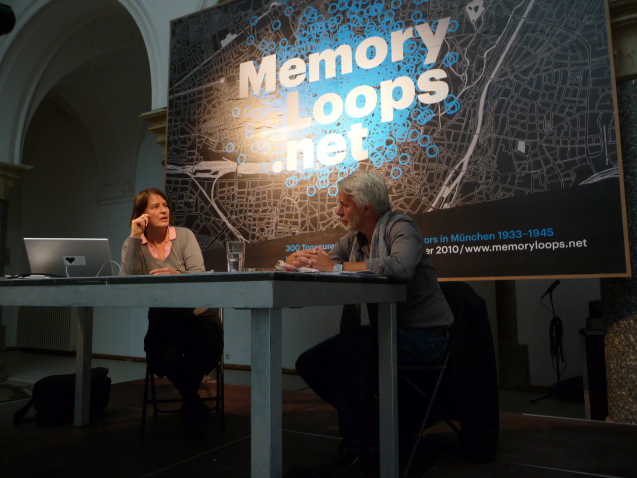 Artist Michaela Melián sits on a podium on the left and curator Chris Dercon on the right. Both face each other in conversation. A large banner of the Memory Loops logo can be seen in the background.