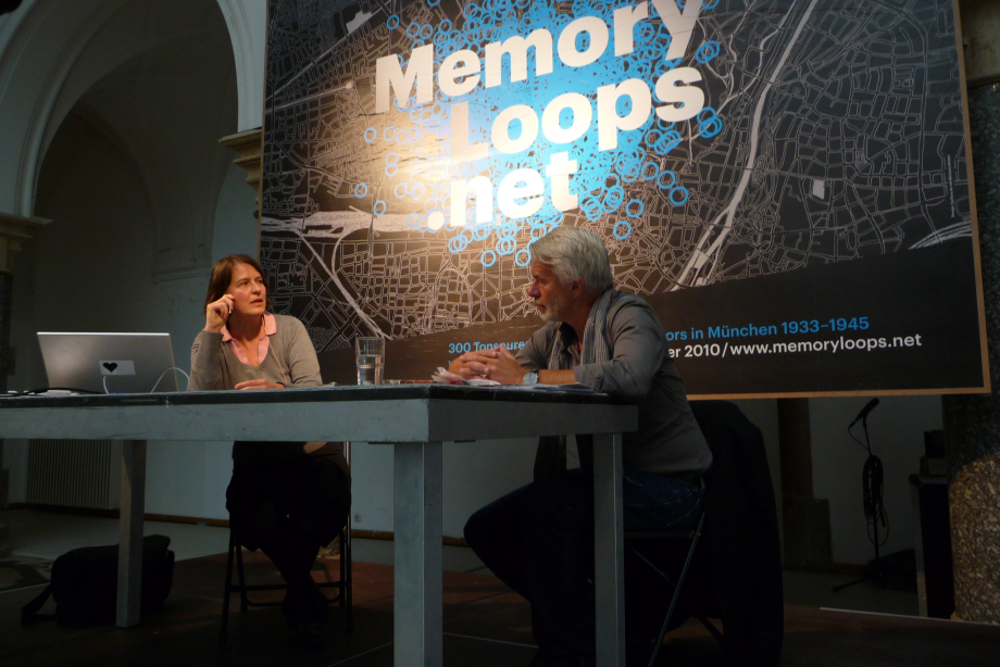 Auf einem Podium sitzen links die Künstlerin Michaela Melián und rechts der Kurator Chris Dercon. Beide sind im Gespräch zueinander zugewandt. Im Hintergrund ist ein großes Banner des Logos der Memory Loops zu sehen.
