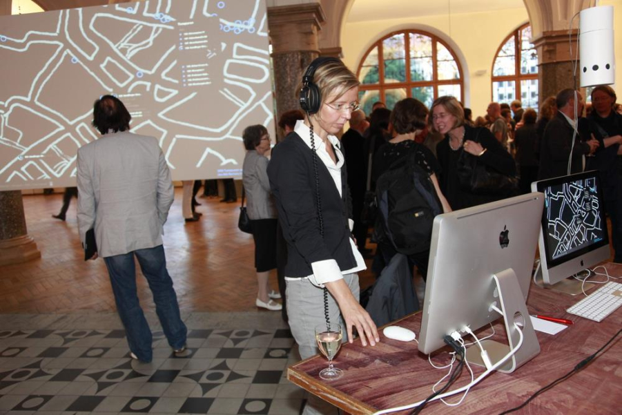 A woman stands in front of a PC with headphones and listens to the soundtracks of the memoryloops. In the background you can see the audience at the opening ceremony and a large screen with the start page of memoryloops.net