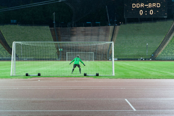 Die Fotografie zeigt das Innere des Olympiastadions bei Nacht. Zu sehen ist der Künstler Massimo Furlan im grün-schwarzen Fußballtrikot in Rückenansicht, der in einem Tor steht. Vor ihm erstreckt sich der grüne Fußballrasen, er ist jedoch der einzige Spieler auf dem Platz. Auf einer Anzeigetafel hoch über den Zuschauerrängen erscheint der Spielstand dieses fiktiven Fußballspiels: "DDR gegen BRD – 0 zu 0".