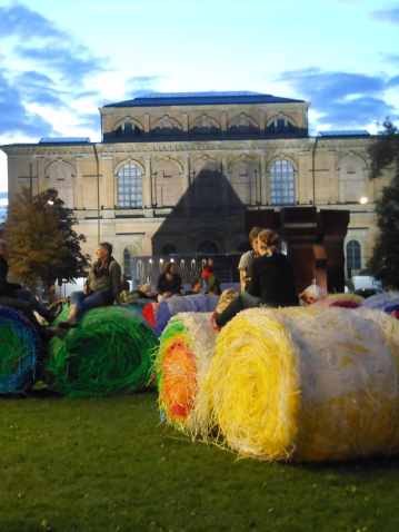 In the evening sun, young people sit relaxed on top of the colored bales and chat. In the background the Alte Pinakothek, illuminated by the summer evening sun.